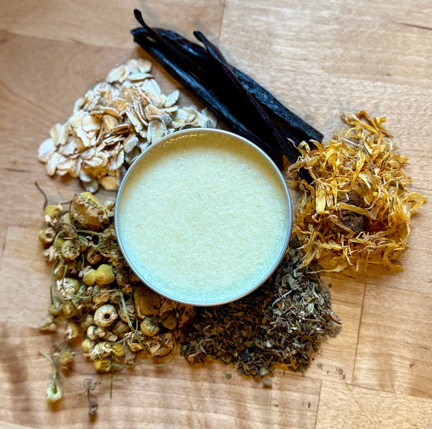 Healing Meadow Salve in tin surrounded by oats, chamomile, calendula, yarrow, and vanilla beans on wooden surface.