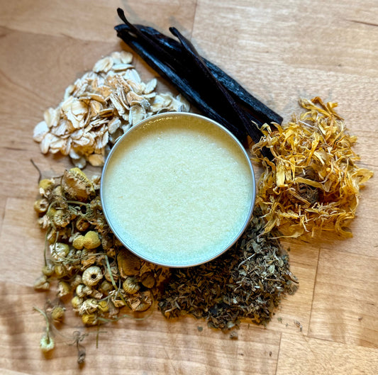 Healing Meadow Salve in tin surrounded by oats, chamomile, calendula, yarrow, and vanilla beans on wooden surface.